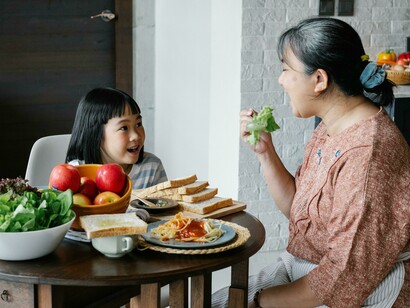 An elderly grandmother sitting at the table eating a healthy salad while her Asian grandchildren watch with delight