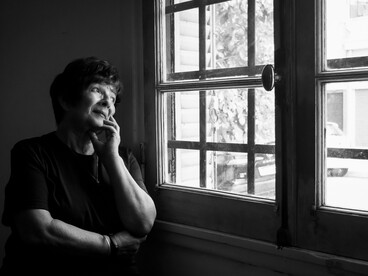 A senior woman looks out of a window in quiet contemplation, her expression conveying grief and remembrance
