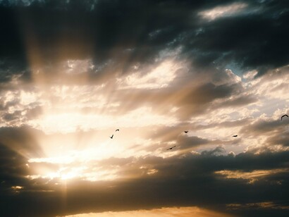Um feixe intenso de luz atravessa as nuvens ao entardecer, enquanto pássaros sobrevoam a paisagem. A revelação pode parecer libertadora, mas enxergar além das aparências também significa abandonar algumas das certezas que sustentam a vida cotidiana