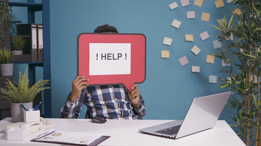 A worker raising a “HELP!” message while seated at a computer, symbolizing how a need for assistance can become an opening for social engineering