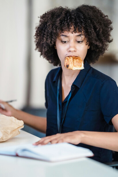 In an office setting, a woman multitasks between paperwork and eating a croissant, illustrating stress and anxiety