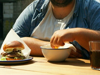 Una persona siede a un tavolo di legno, mangiando un grosso hamburger e patatine: "Si può descrivere la condizione dell’informazione odierna ricorrendo all’esempio del fast-food. L’attrattiva del fast-food risiede in due elementi: costa poco ed è veloce" 