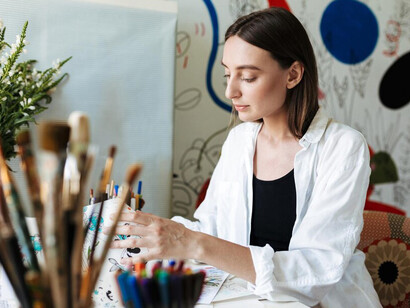 A girl holds paintbrushes in her hand, gazing dreamily at the camera, with a large patterned canvas in the background, capturing the artistic workspace and creative process at home