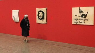 Visitante observa obras de Hélio Oiticica em exposição, entre elas a emblemática "Seja marginal seja herói". Inspirada na morte de Manuel Moreira, conhecido como "Cara de Cavalo", a criação tornou-se um manifesto artístico contra a violência e a exclusão, reafirmando a capacidade da arte de desafiar convenções — movimento que também marcou a literatura brasileira a partir da Geração de 45. Ao transformar um episódio de violência em reflexão artística, Oiticica ajudou a redefinir os caminhos da arte contemporânea brasileira, em sintonia com a renovação da literatura e de outras formas de expressão