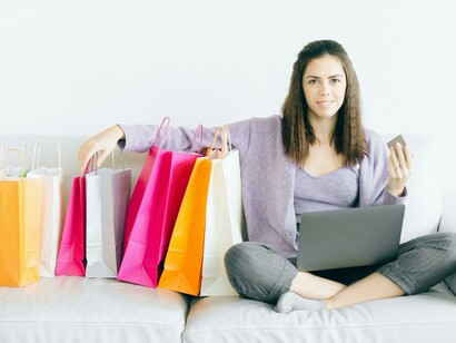 A woman sits on a couch with paper shopping bags piled around her, reflecting the theme of shopping addiction
