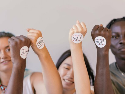 Mãos de diferentes tonalidades de pele se erguem com adesivos de votação em um gesto de solidariedade. A imagem amplia a ideia de escolha individual para uma democracia que precisa reconhecer e proteger diferentes grupos sociais