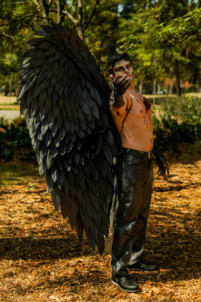 A shirtless man in an angel costume holding a bow, embodying a fallen-angel aesthetic with symbolic wings