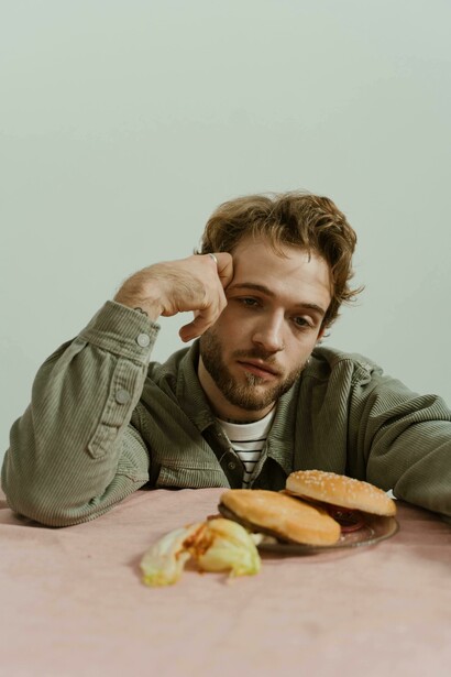 A man sits in a minimalist studio, holding his head as he looks at a burger on the table, reflecting a depressed mood