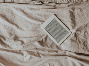 Um leitor de livros digitais repousa sobre uma cama cuidadosamente arrumada, cercado pela textura suave dos lençóis e travesseiros. O livro muda de suporte, mas permanece presente como companhia para momentos de pausa e intimidade