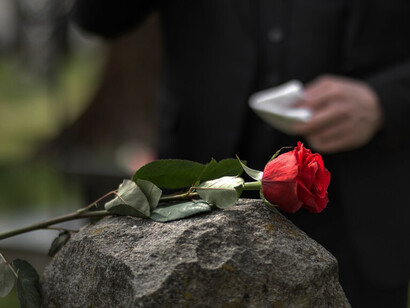 A gravestone with fresh flowers rests in the cemetery, while mourners gather for a burial ceremony and a funeral procession moves slowly nearby