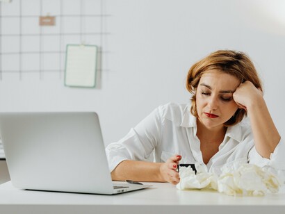 A woman sitting in front of her laptop, surrounded by crumpled papers, representing the paralysis and frustration caused by self-doubt