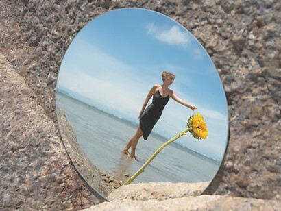 A woman, radiating summer vibes, strikes a pose at the beach, with a round mirror reflecting her image, creating a visually captivating and atmospheric moment