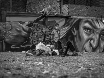 On the street, a homeless man lies on a sofa, with striking graffiti art covering the wall behind him