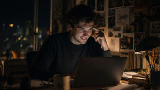 A young man sits at a dimly lit desk, smiling softly as he looks at his laptop, surrounded by scattered papers and warm ambient light