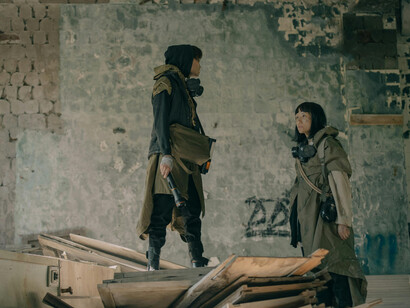 A man and a woman stand together inside a war-torn ruin, surrounded by crumbling walls and silence