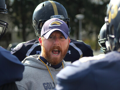 A coach in a blue and white jacket, wearing a black helmet, shouts instructions during an American football training sessio