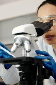 A woman wearing gloves and a lab coat looking at a sample through a microscope