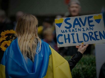 A woman draped in the Ukrainian flag holding a sign that reads "save Ukrainian children"