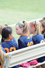Female football players sitting on a bench during a daytime match, resting between plays