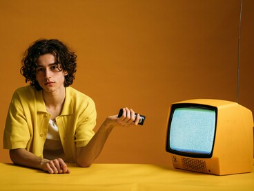 Com um comando remoto na mão, o jovem olha para a câmara enquanto, ao seu lado, o televisor exibe uma tela coberta de interferências. A cena sugere a facilidade de mudar de canal e de conteúdo, mas também convida a refletir sobre a diferença entre controlar o que se vê e compreender criticamente a informação recebida