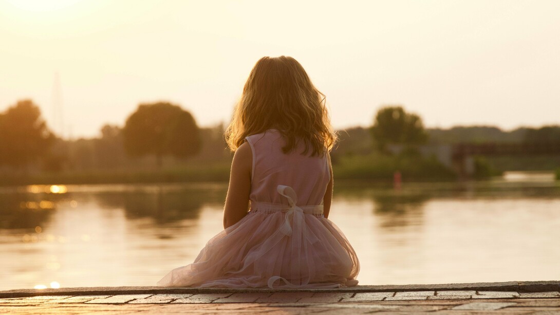 A young girl sitting alone beside the water at sunset, representing the loneliness that can begin with the fear of being left