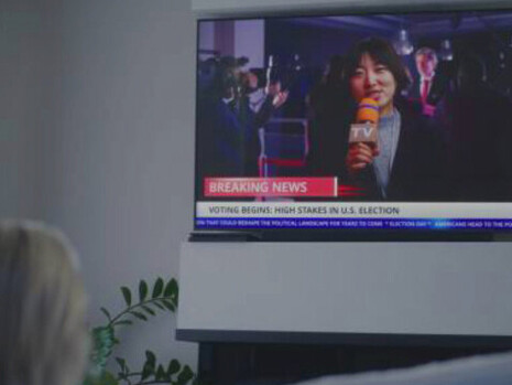 A woman sits on the couch in her living room, watching a live news broadcast, feeling overwhelmed by the constant stream of political updates