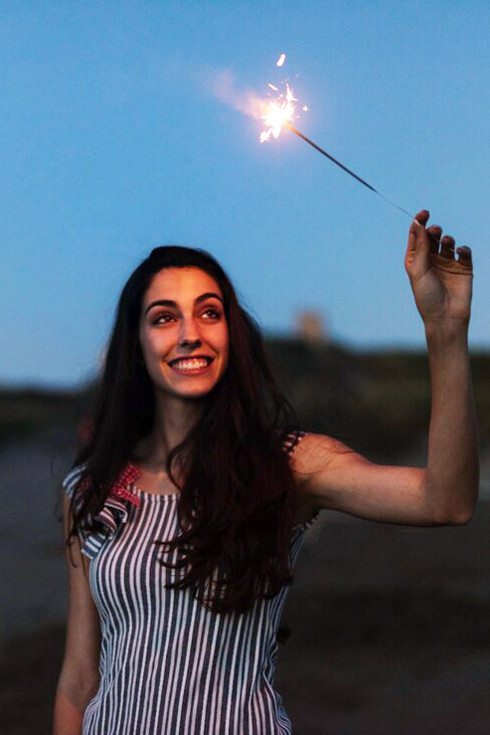 A woman standing on a beach at night, holding a sparkler with sparks of energy flowing from her fingertips