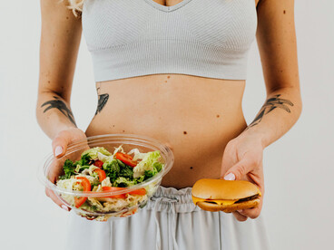 A woman in a white tank top holds a bowl of vegetable salad in one hand and a hamburger in the other