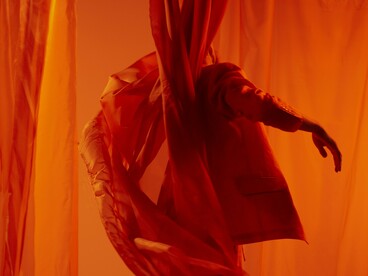 Entre cortinas vermelhas, um homem dança enquanto o tecido acompanha o movimento do corpo. O palco torna visível aquilo que o texto chama de fogo interior: uma energia que pede para ser expressa. Vermelho sobre vermelho, corpo e tecido parecem compartilhar o mesmo impulso. Há uma chama que não precisa ser vista para existir; basta encontrar uma forma de movimento