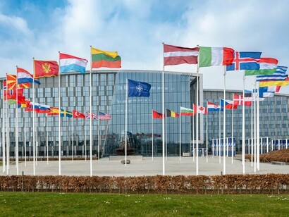 Bandeiras dos países-membros tremulam em frente à sede da OTAN, uma das principais instituições criadas para preservar a segurança coletiva no cenário internacional. O artigo analisa como as transformações recentes na política externa dos Estados Unidos colocam em debate a estabilidade da ordem geopolítica construída após a Guerra Fria. Sede oficial da OTAN em Boulevard Leopold III, 1110 Bruxelas, Bélgica