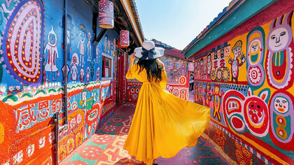 A woman stands in Rainbow Village in Taichung, Taiwan, wearing a long yellow dress, surrounded by vibrant, colorful street art and murals