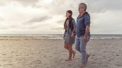 Pai e filha caminham lado a lado à beira-mar. Relações de afeto e cuidado também moldam a forma como homens envelhecem. Entre passos lentos e companhia constante, o cuidado aparece como vínculo e não apenas obrigação. Em muitos casos, são justamente as mulheres que sustentam redes emocionais e de cuidado ao longo da vida. O texto discute como masculinidade, afeto e cuidado influenciam até a expectativa de vida