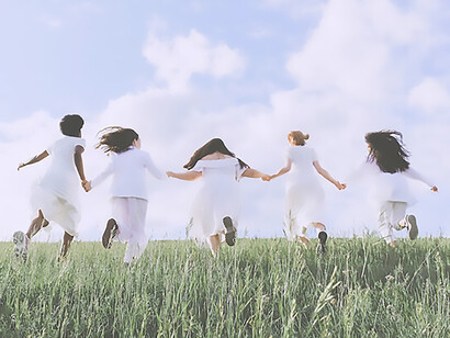 Five women, adorned in flowing white dresses, gracefully traverse the lush green landscape hand in hand, embracing the beauty of nature
