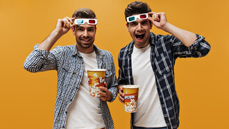 Two men in white T-shirts and blue checkered shirts, holding popcorn and removing 3D glasses, watching a movie against an isolated orange background