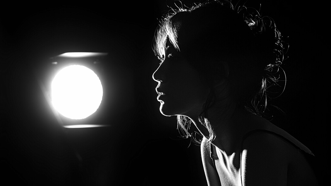 A silhueta de uma mulher emerge diante de uma única luz, enquanto o resto permanece em sombra. Em “Joshua”, Elsa Mangue transforma esse contraste entre escuridão e claridade numa recusa que também é sobrevivência. A imagem ecoa a força íntima de quem fecha a porta para não morrer por dentro