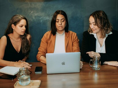 Three women gathered around a laptop in discussion, representing collaboration, shared responsibility, and the human dimension of modern work