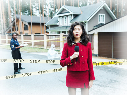 Standing in front of police tape at the scene of a crime and a policeman writing their report, a female news anchor waits to report the news