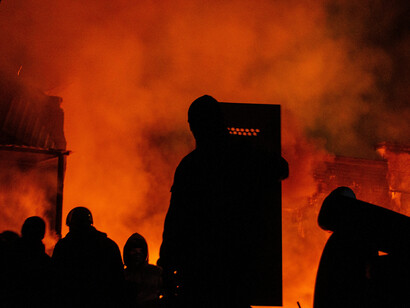 A crowd of silhouetted figures stands in a dark field, positioned behind police barriers under the night sky