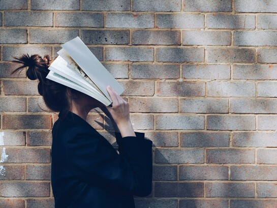 A schoolgirl stands against a rustic red brick wall, holding a book up to cover her face, reflecting student burnout