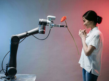 A robot holds out a flower, offering it tenderly to a woman wearing a white T-shirt