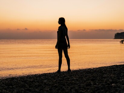 Donna in riva al mare al tramonto: “Mi piace sedermi sulla riva al tramonto, lasciando che la luce dorata del sole si rifletta sull’acqua. In quei momenti, il tempo sembra rallentare, permettendomi di godere di una pace rara e preziosa” 