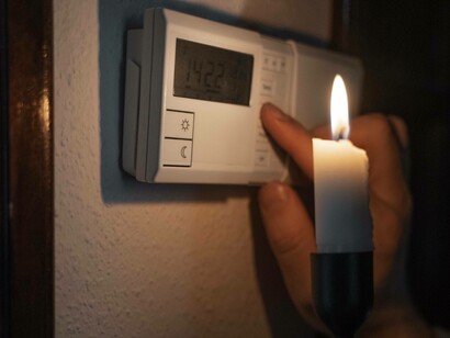 Una mano che regola un termostato digitale su una parete, illuminato dalla fiamma di una candela, durante un'interruzione di corrente