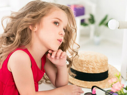 A young girl sitting by the mirror in her mother’s bedroom, playing with cosmetics, reflects issues related to children’s exposure to beauty products, the impact of makeup on young girls, and childhood self-image challenges
