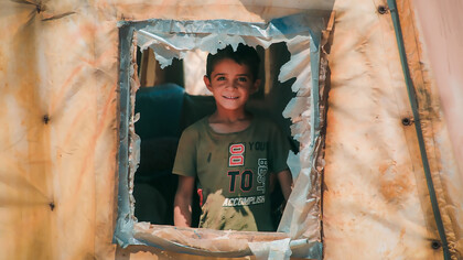 A young boy gazing out from the window of a refugee camp tent, symbolizing the hardship and uncertainty faced by displaced Syrian families