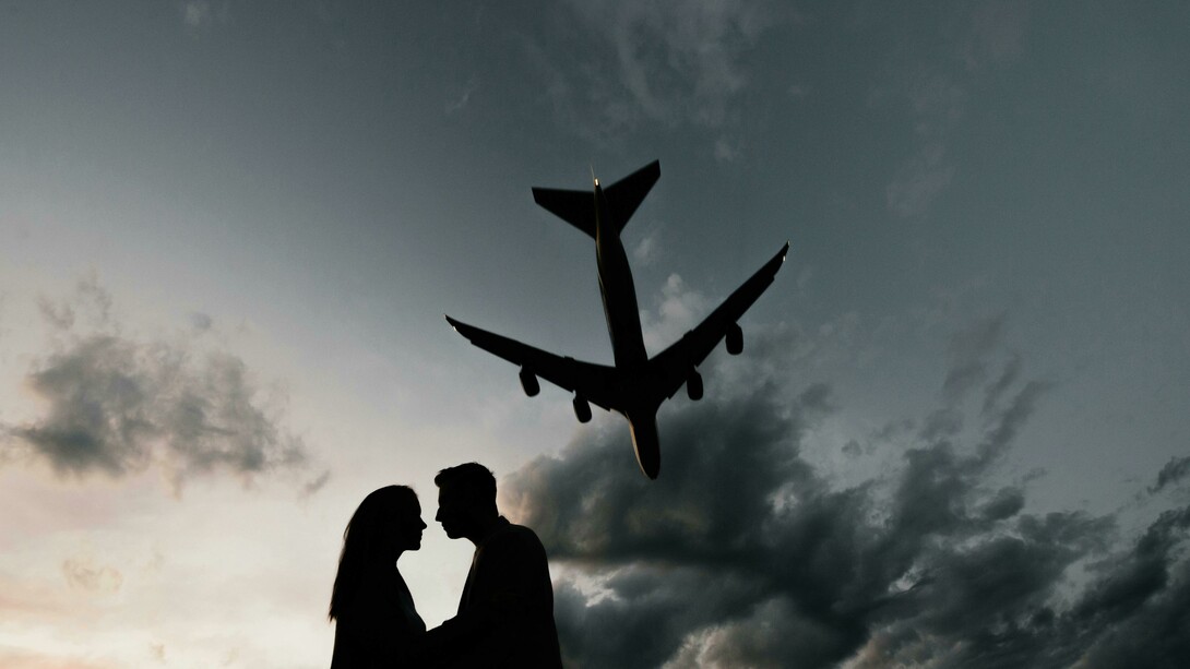 Silhouettes of a couple sharing a farewell embrace under a plane flying across the sky at the airport