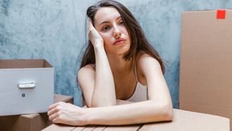 A woman, encircled by boxes, wears a somber expression, reflecting her emotions as she prepares for a move