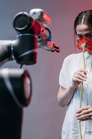 A woman delicately cradles a vibrant red flower while accompanied by an AI-driven robot