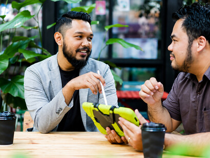 Two men sit in a café surrounded by lush greenery, sharing coffee and discussing their personal challenges