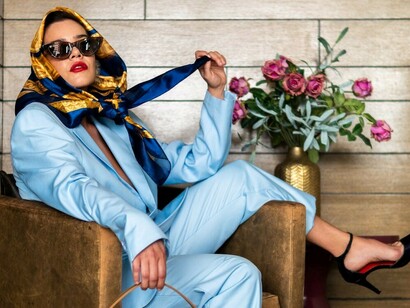 An elegant woman sits gracefully, dressed in a light blue suit paired with a chic headscarf, stylish sunglasses, and classic black heels