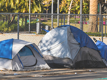 A row of makeshift homeless shelters positioned in front of upscale residential buildings, highlighting the contrast between affluence and urban poverty